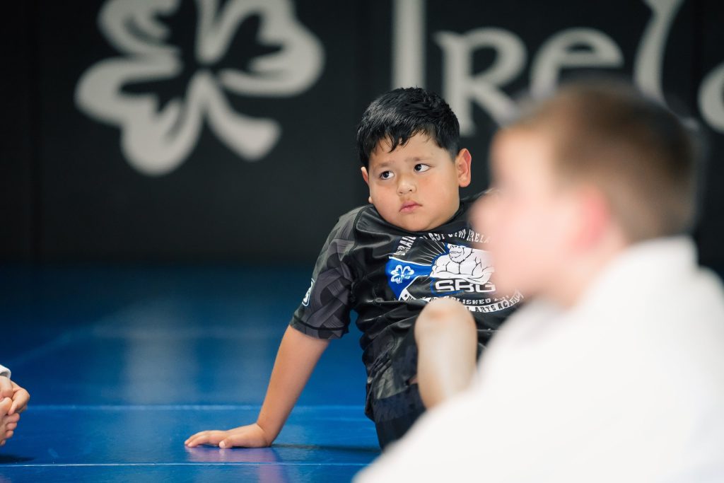 Boy in an SBG rash guard sitting on the blue mats during an SBG Kids class